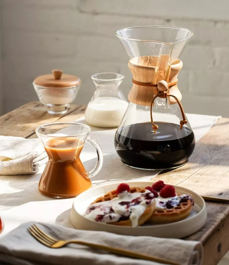 Una cafetera casera con una taza de café y gofres sobre una mesa.